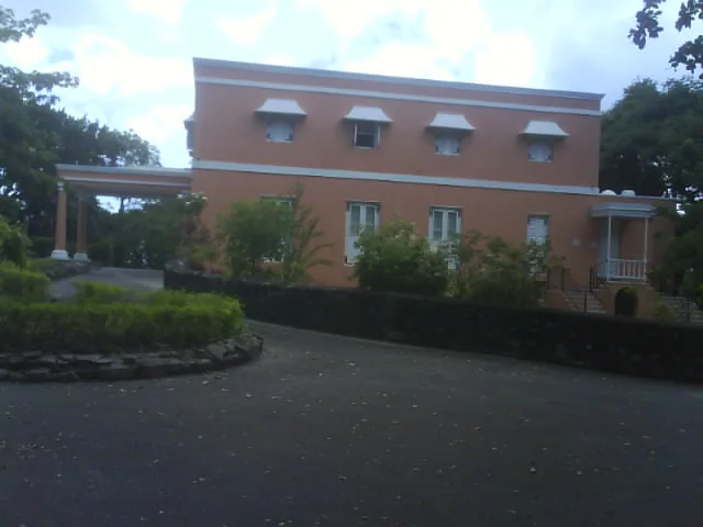 Wildey Great House in St Michael, Barbados, seen from the drive: a long two-storey house with thick walls, a low roof line behind a parapet, shuttered upper openings and a colonnaded porch