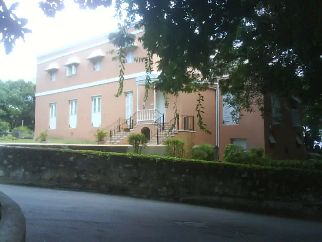 Side view of Wildey Great House, Barbados, framed by trees, with a coral stone boundary wall in the foreground and shuttered windows along the elevation